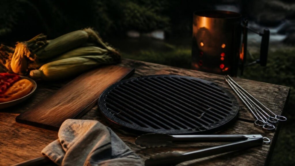 Cast iron grill grate and stainless steel tongs and skewers on a wood table at dusk, with corn in the husk and a lit charcoal chimney behind